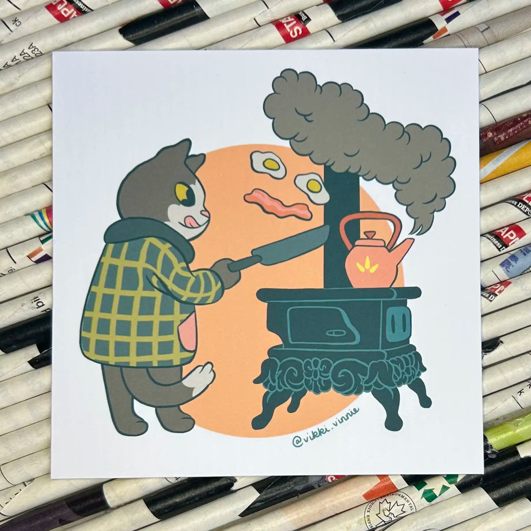 An art print designed by Vikki Vinnie depicting a cat cooking breakfast on a wood stove.
