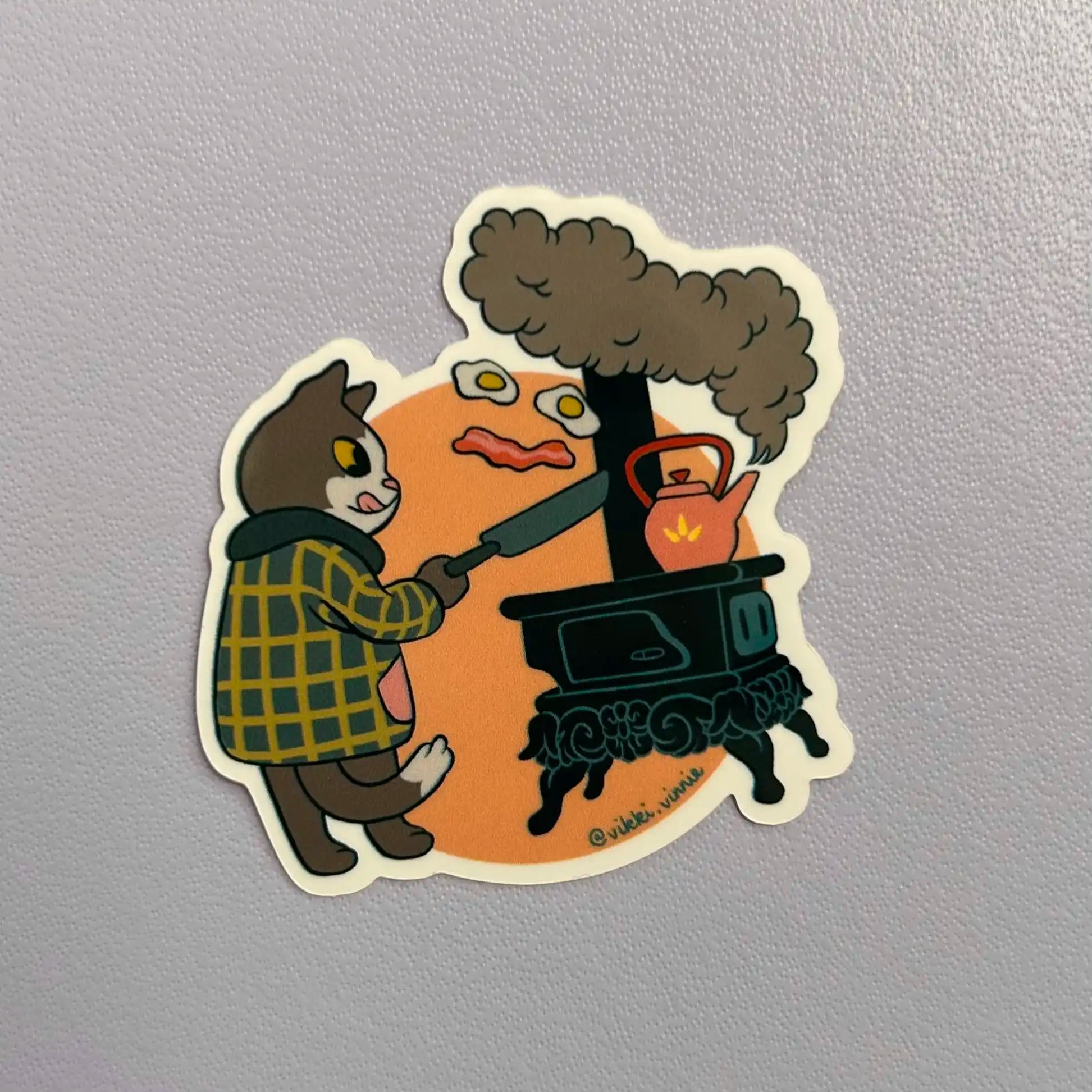 A die cut vinyl sticker designed by Vikki Vinnie depicting a cat cooking breakfast on a wood stove