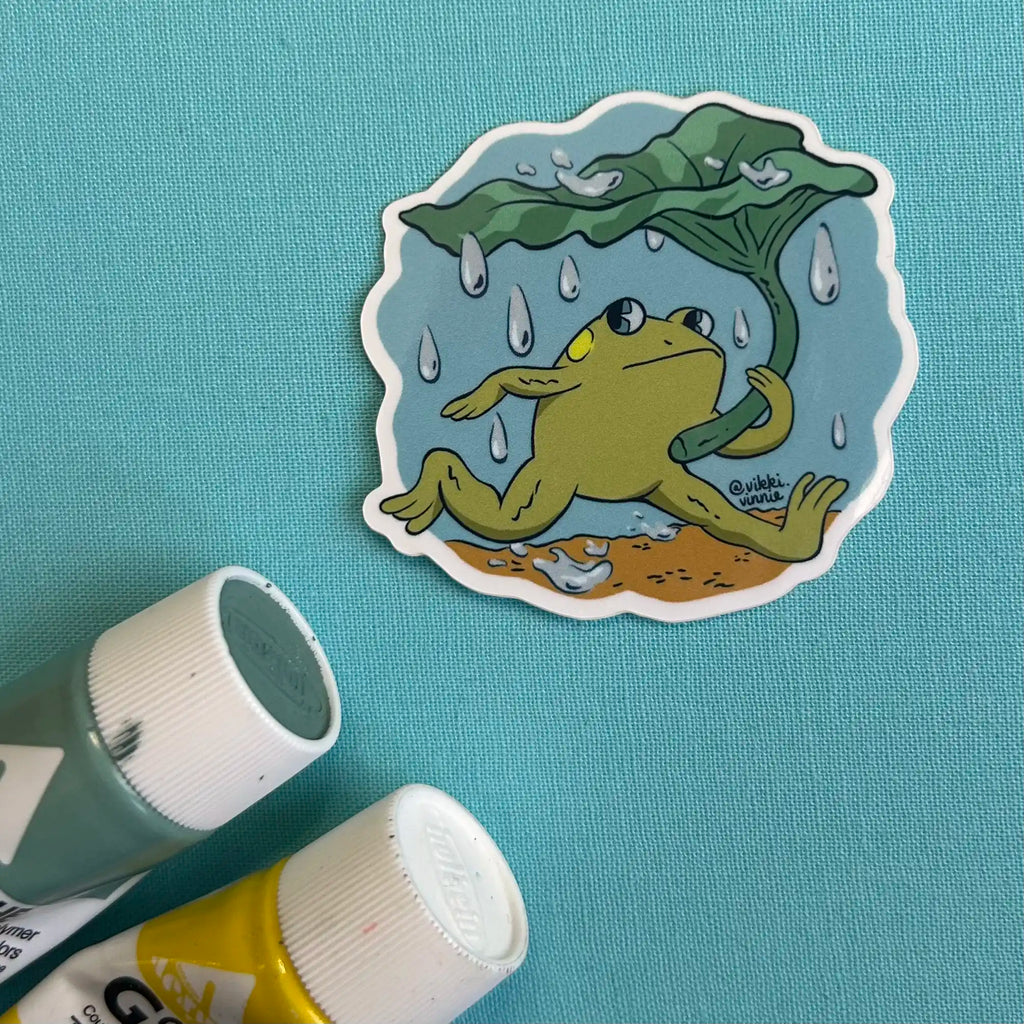 A vinyl die cut sticker designed by Vikki Vinnie depicting a frog using and leaf as an umbrella during a rainstorm