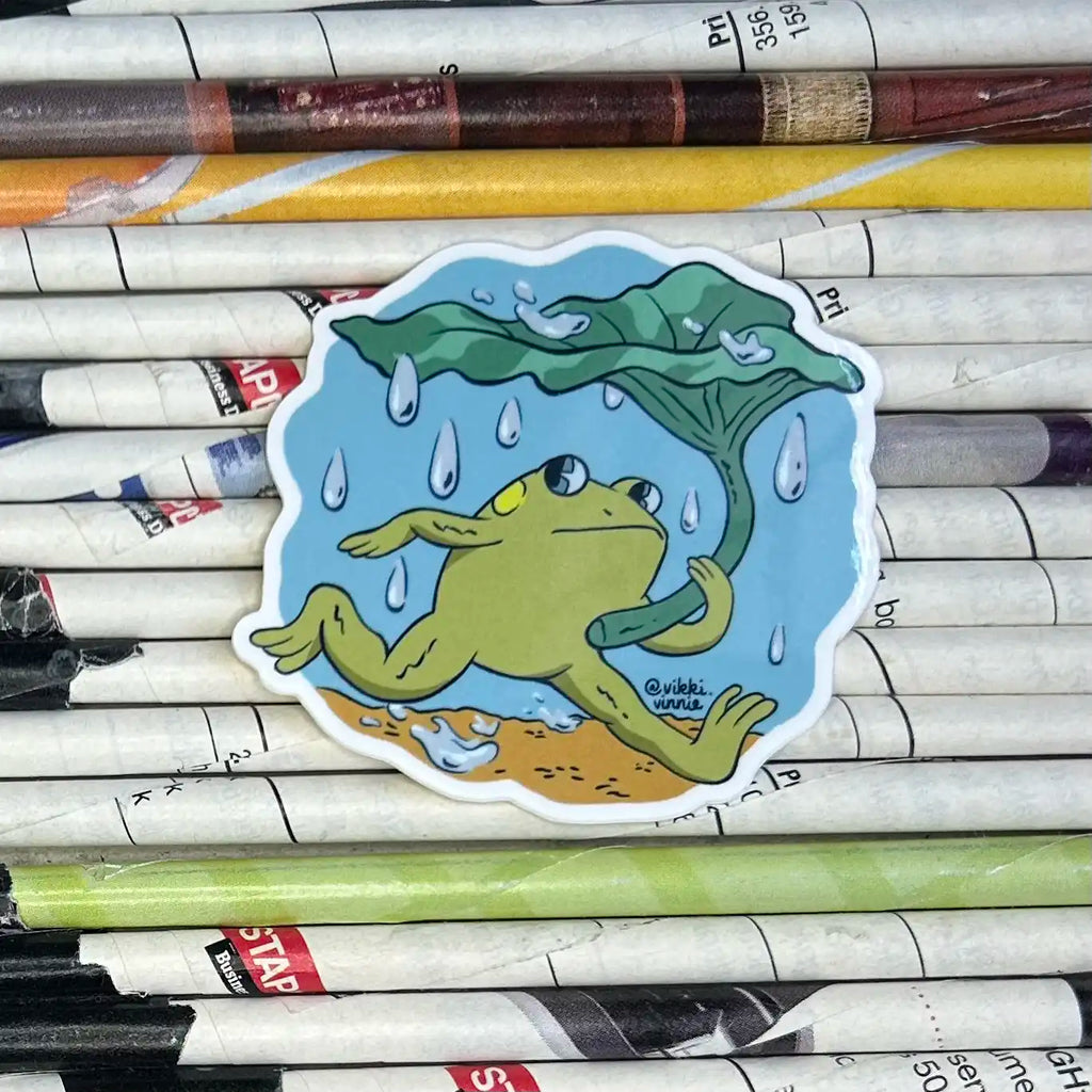 A vinyl die cut sticker designed by Vikki Vinnie depicting a frog using and leaf as an umbrella during a rainstorm